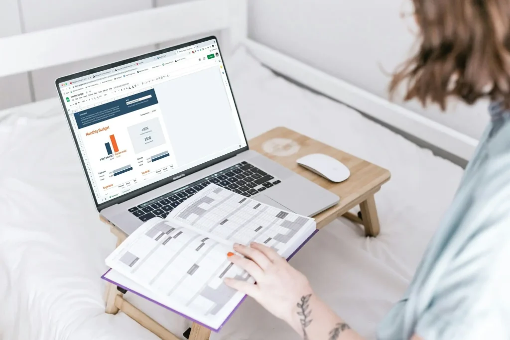 a person sitting on a bed with a laptop and worksheet with budget